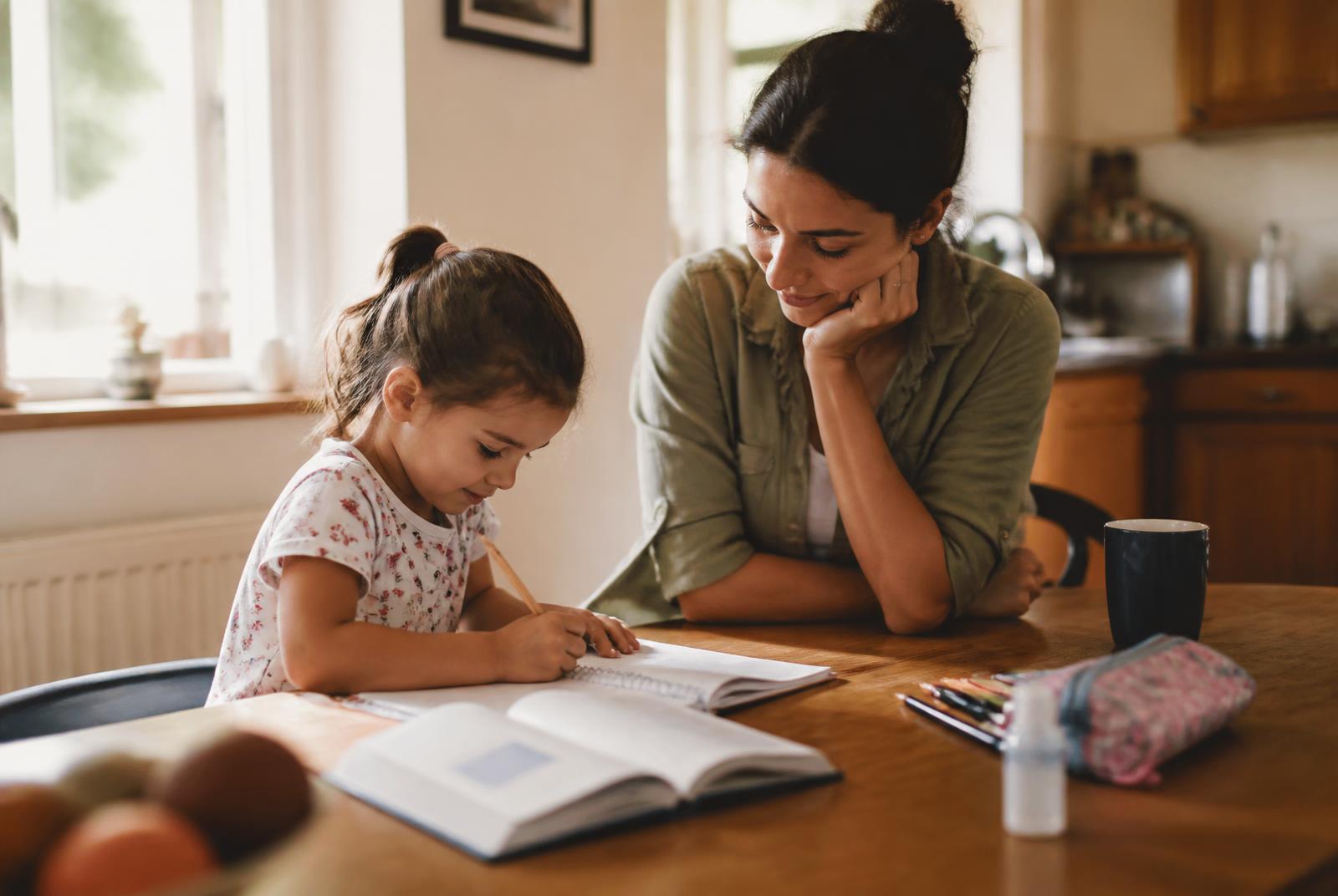 A mother sitting beside her young child at a wooden table doing schoolwork in warm morning light