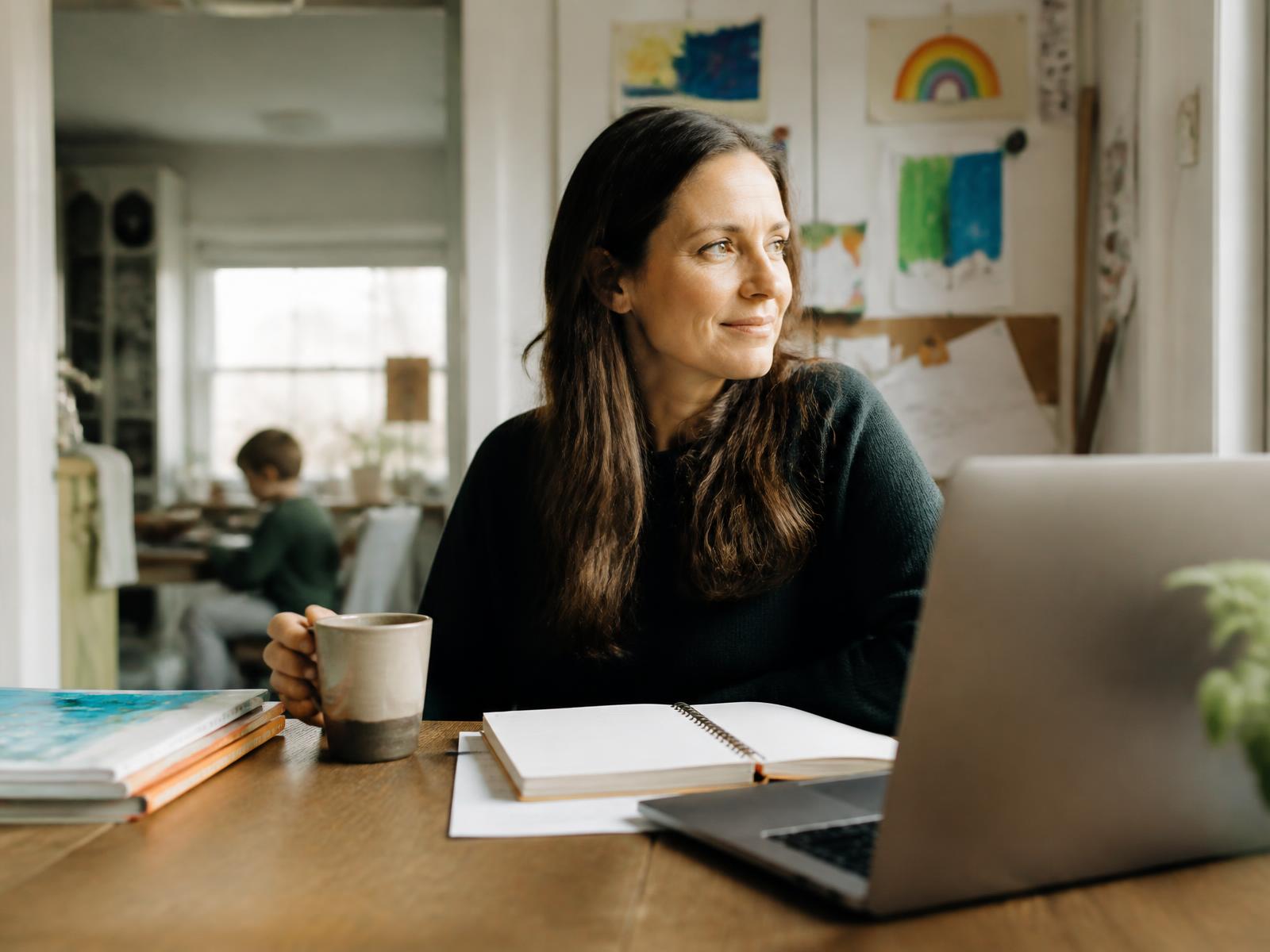 A homeschool mom sitting at a wooden kitchen table with coffee, a notebook, books, and a laptop in warm morning light